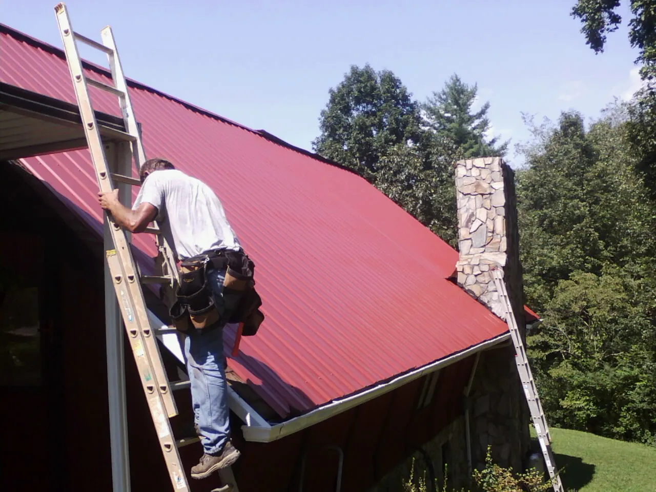 Roofer on a ladder beside a red metal roof and stone chimney — Soffit & Fascia Repair by Roofing Brothers in Erwin